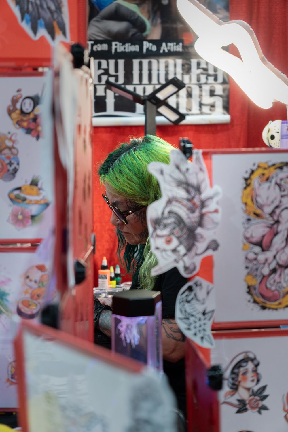 A tattoo artist with green hair works at a decorated booth at a tattoo convention surrounded by flash art designs.