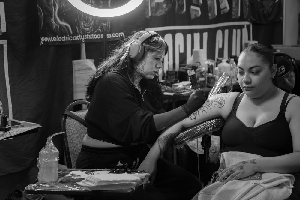 Black and white photo of a tattoo artist working on a client's arm in a tattoo studio.