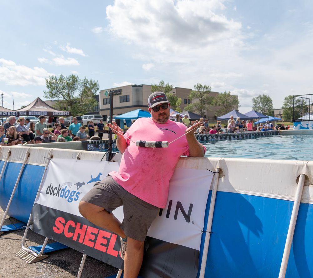 Man in pink shirt posing at Dock Dogs competition pool with Scheels sponsorship banner at outdoor event.