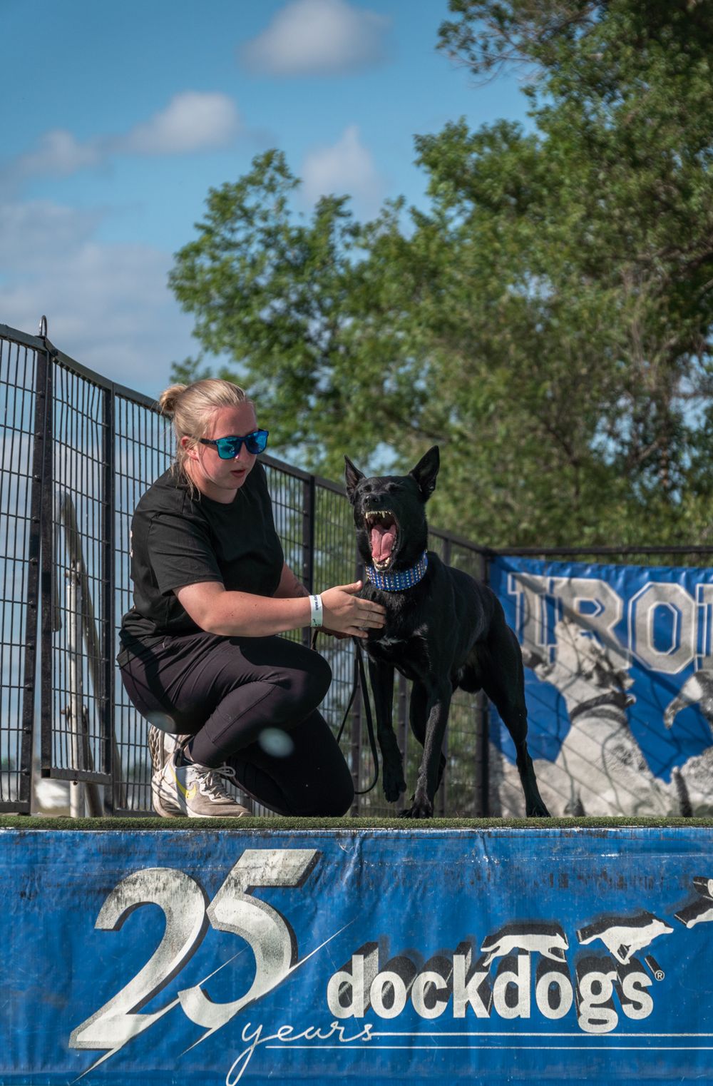 A person in black clothing sits with a black dog on a blue graffiti wall during a photo session outdoors.