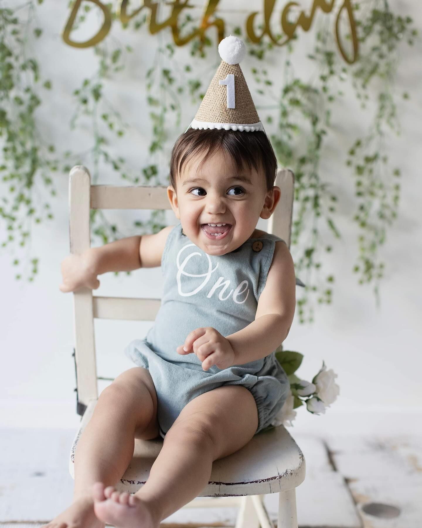 Baby in light blue outfit sits on white chair against floral backdrop during birthday photo session.