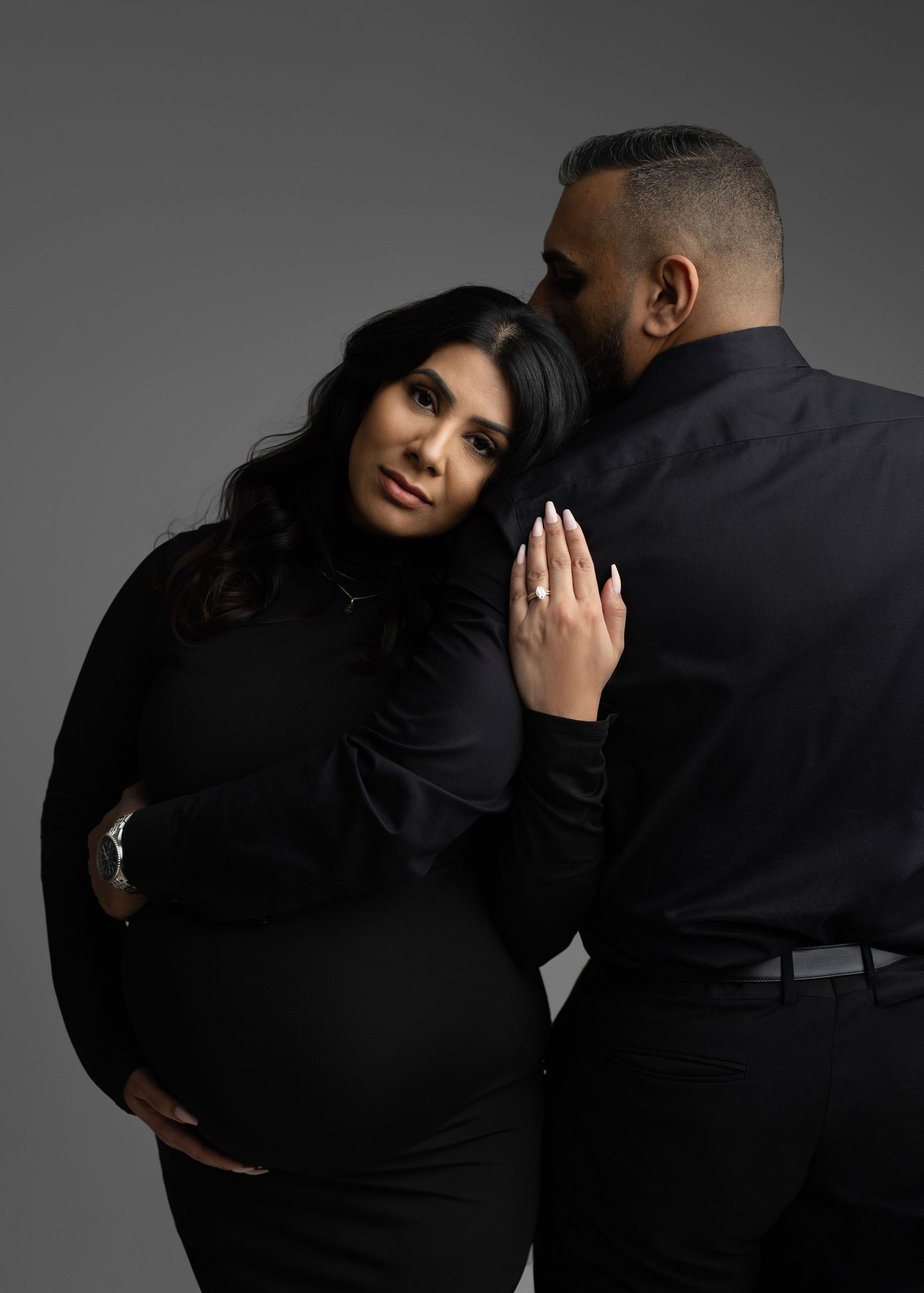 Two people in black clothing embrace and dance together against a gray studio background in a dramatic pose.
