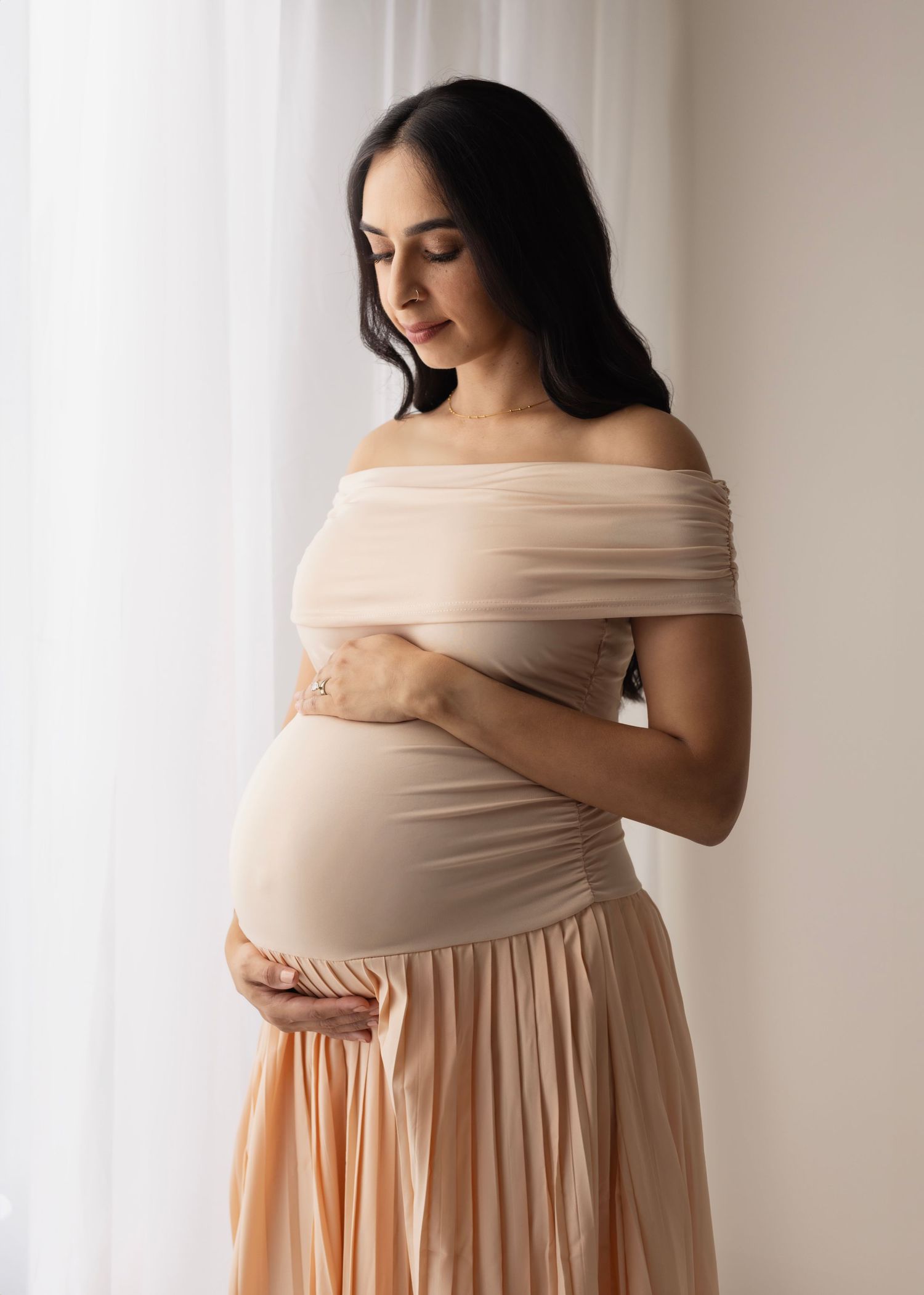 Maternity photo series showing woman in beige off-shoulder dress against white backdrop in four elegant poses.