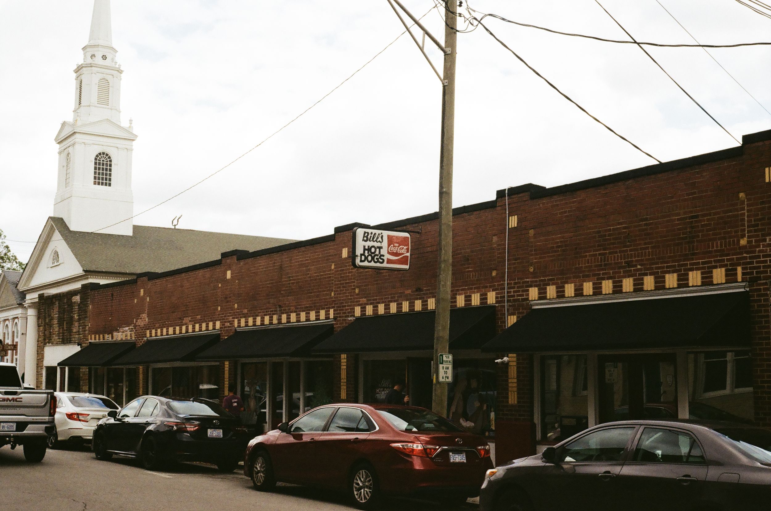Historic brick buildings with storefronts and parked cars line a commercial street in an old downtown district. Bills hotdogs