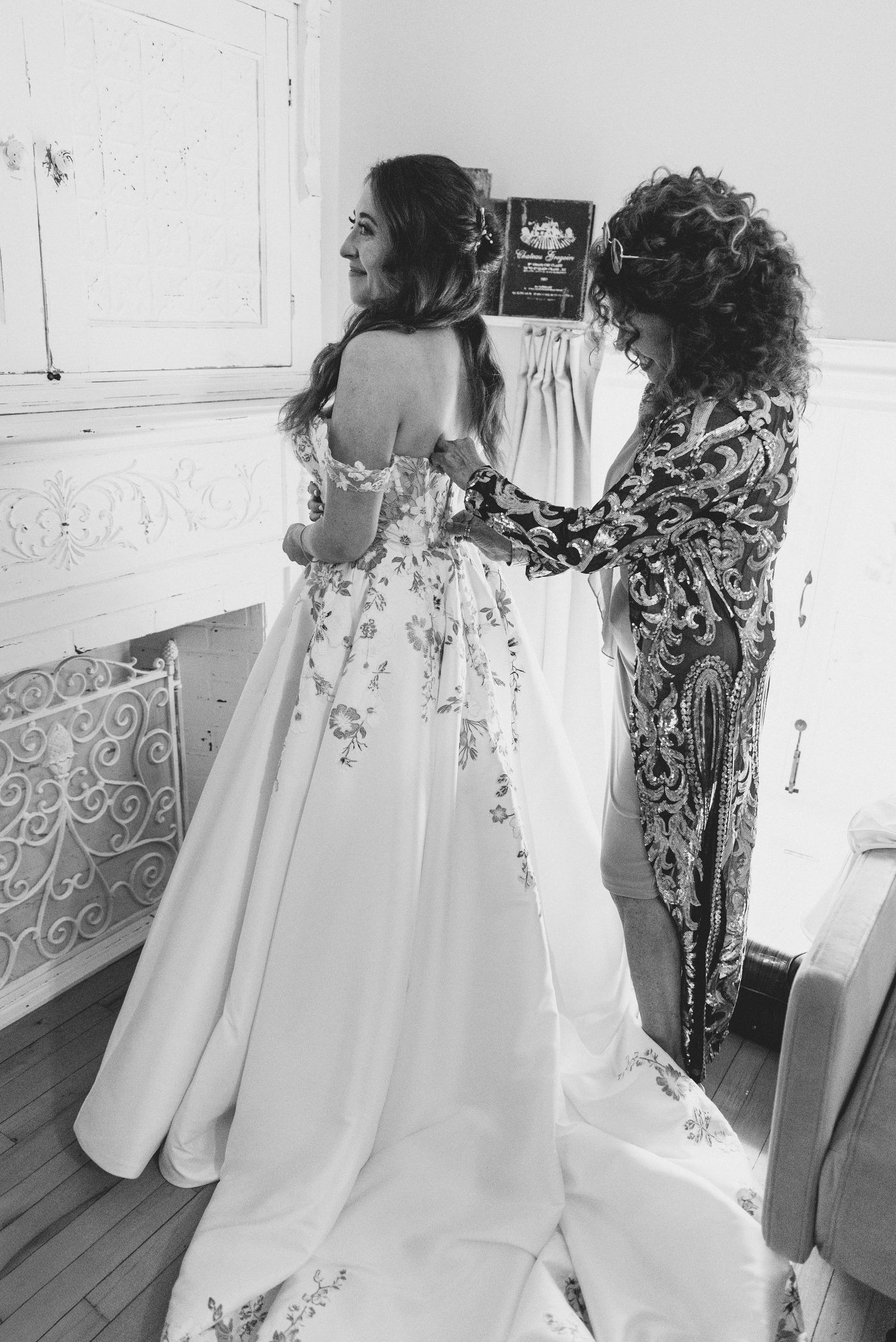 A bride in a flowing white wedding dress with floral embellishments stands next to another person in black and white attire.