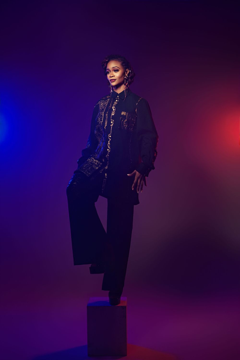 A figure in a dark outfit poses dramatically against a purple and red gradient backdrop in a fashion photoshoot.