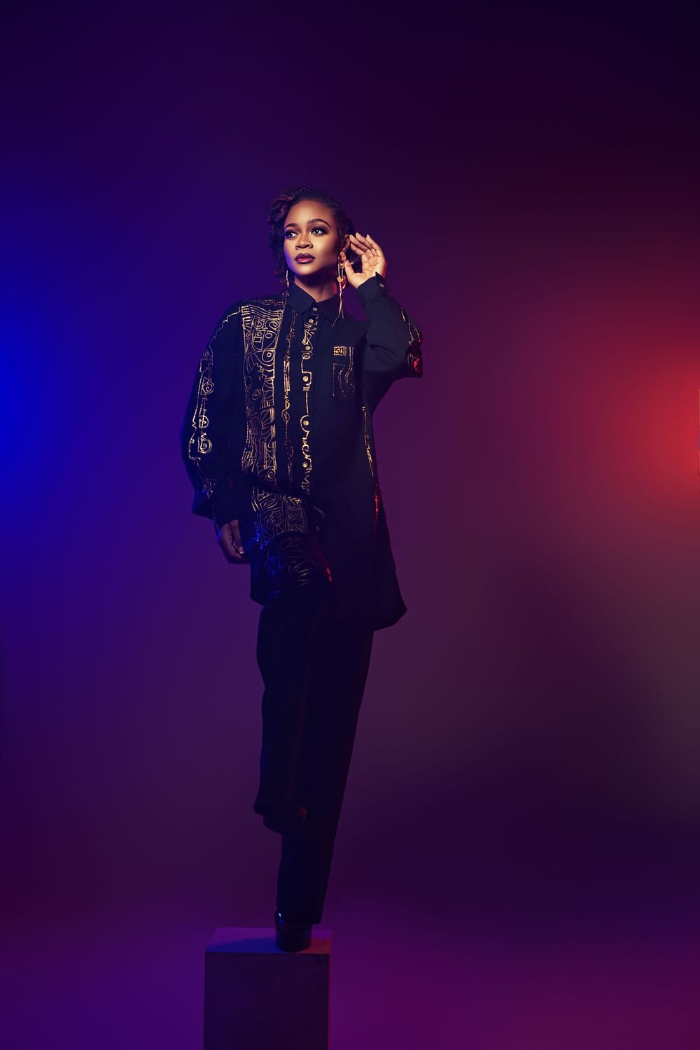 Performer in a black sequined outfit poses dramatically against a purple and blue gradient backdrop on stage.