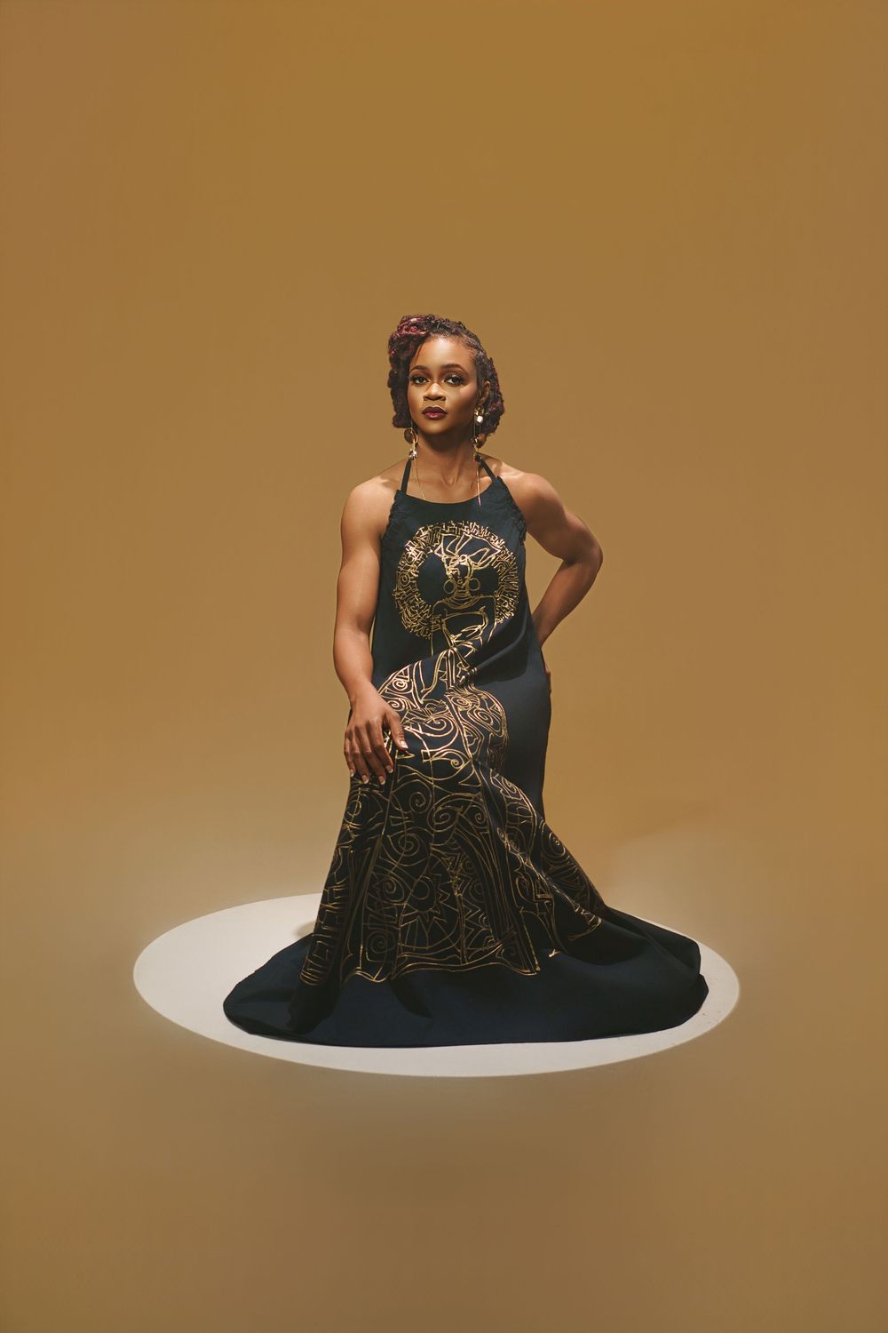 Fashion model poses in elegant black dress with gold embellishments against brown backdrop.