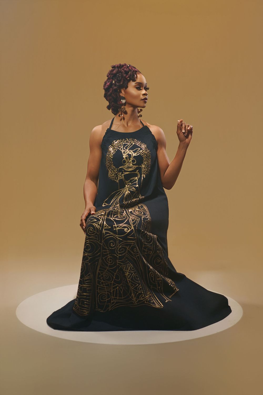Model poses on white platform wearing elegant black dress with gold pattern against warm brown background.