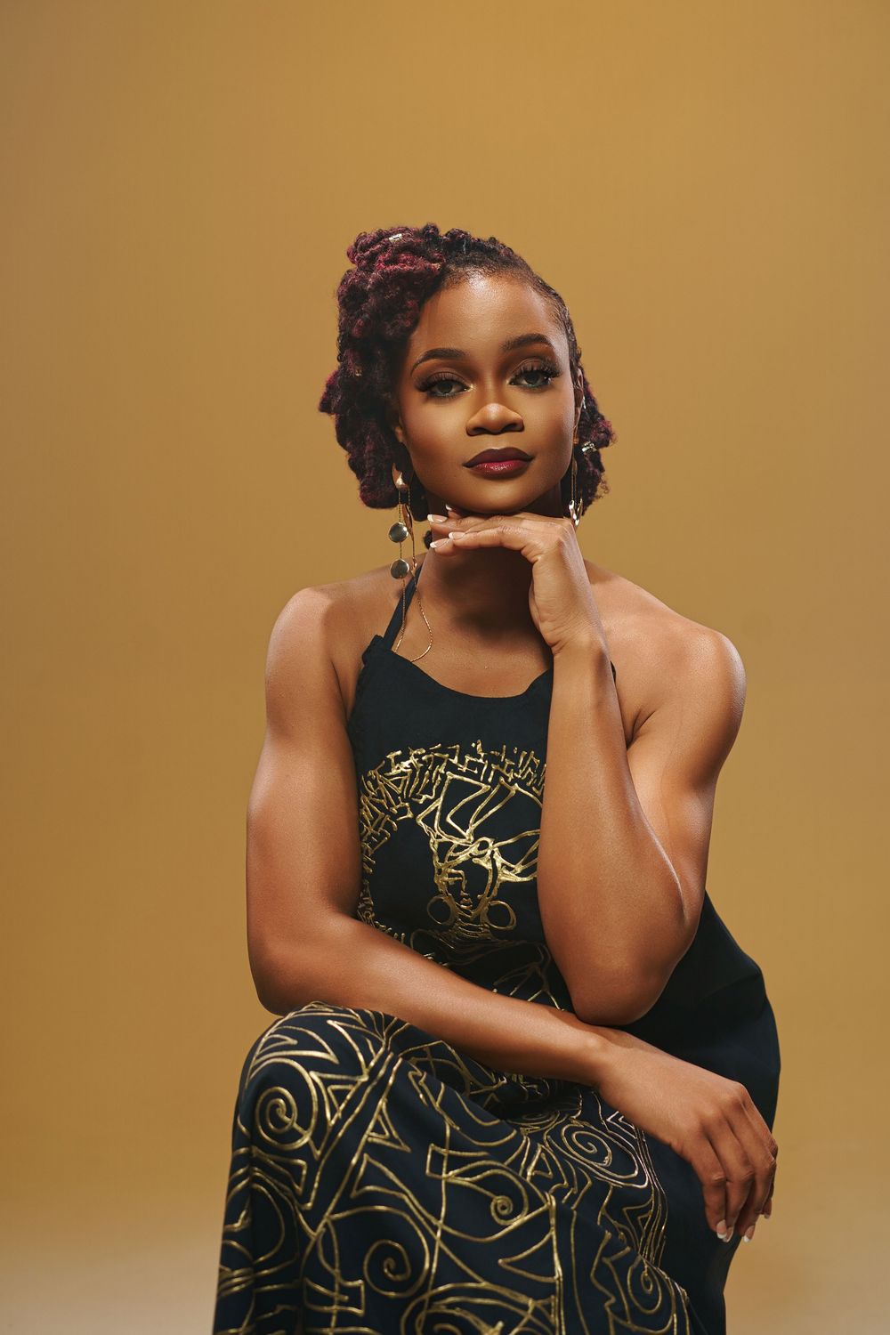 Fashion model poses in elegant black dress with gold geometric patterns against warm brown background.