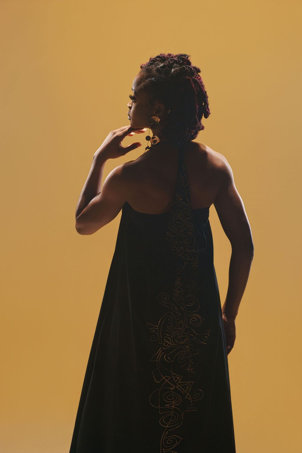 Artistic silhouette shot of model wearing black dress with metallic details against golden backdrop.
