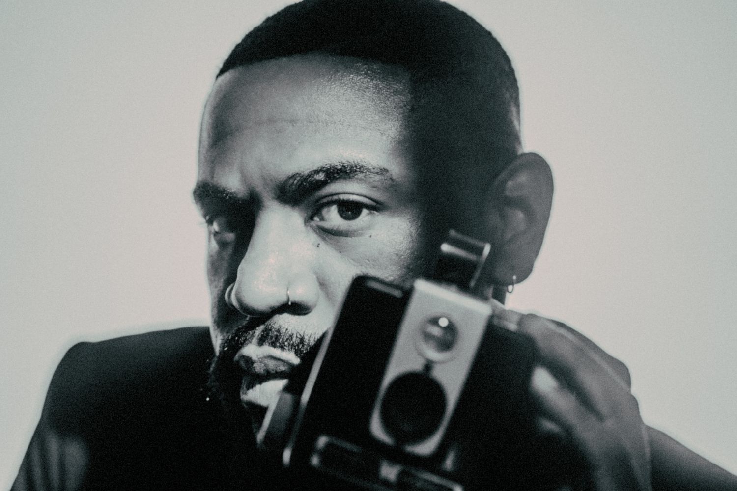 A black and white photograph shows a person holding a camera while looking intently at the viewer.
