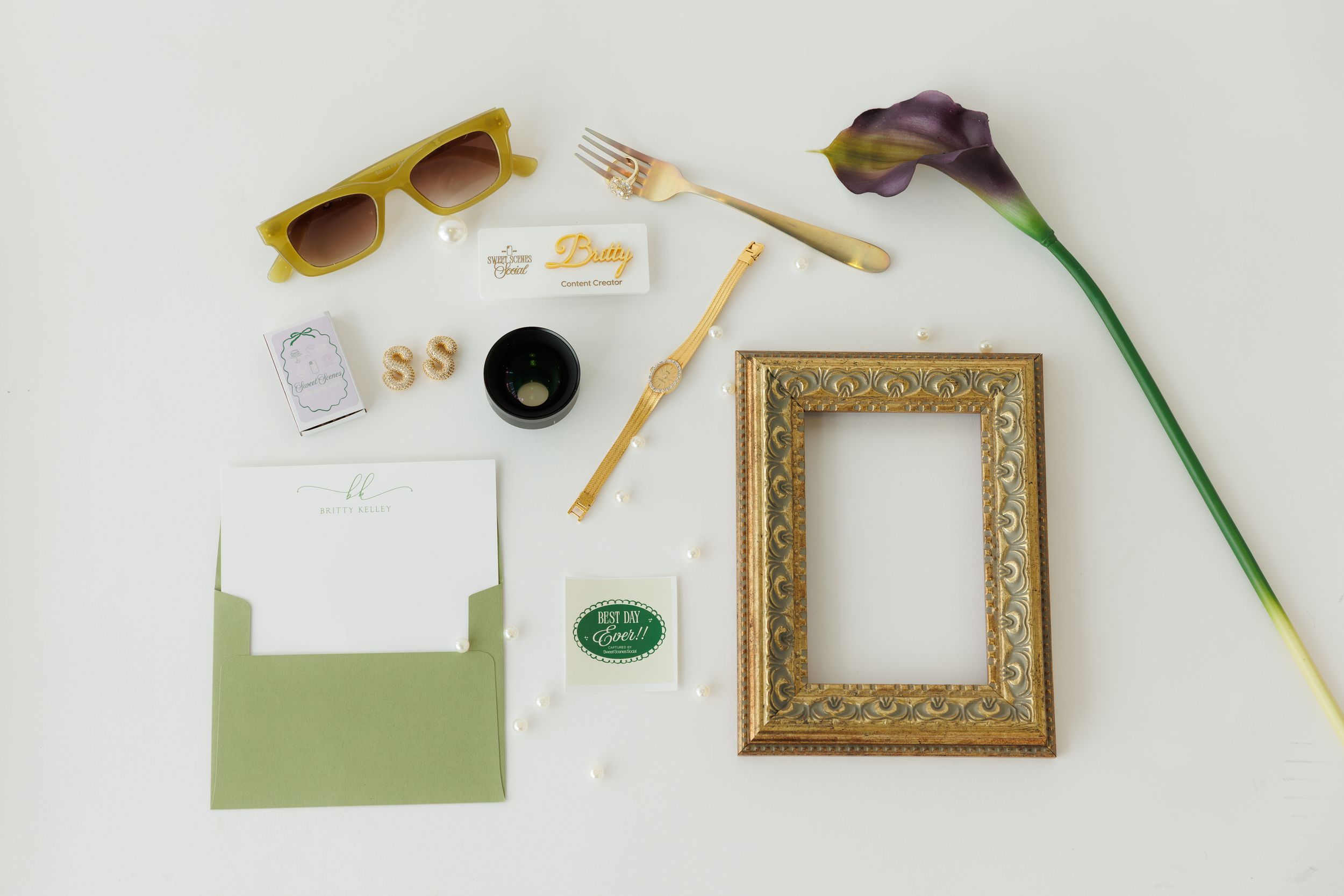 Flatlay of stationery items including sunglasses, gold frame, purple flower, envelopes and writing tools on white surface.