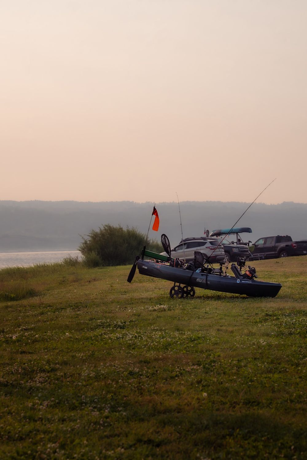 A small fishing boat sits on grassy shoreline at dusk with parked cars and water visible in the background.