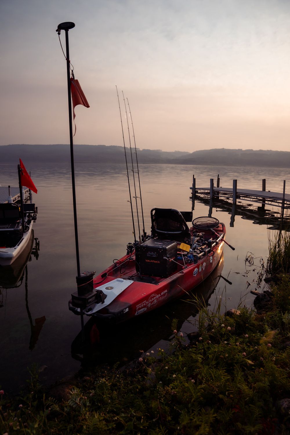 A fishing boat with multiple rods is docked at a tranquil lake pier during sunset with mountains in the background.