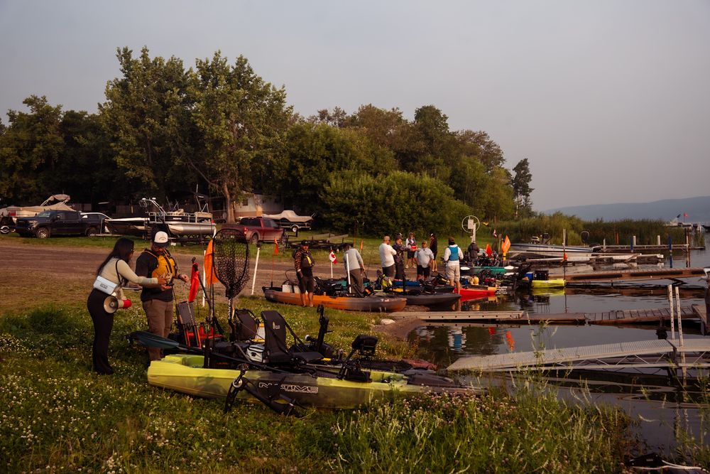 Kayakers and boaters gather at a dock during sunset, with boats and equipment lined up along the grassy shoreline.