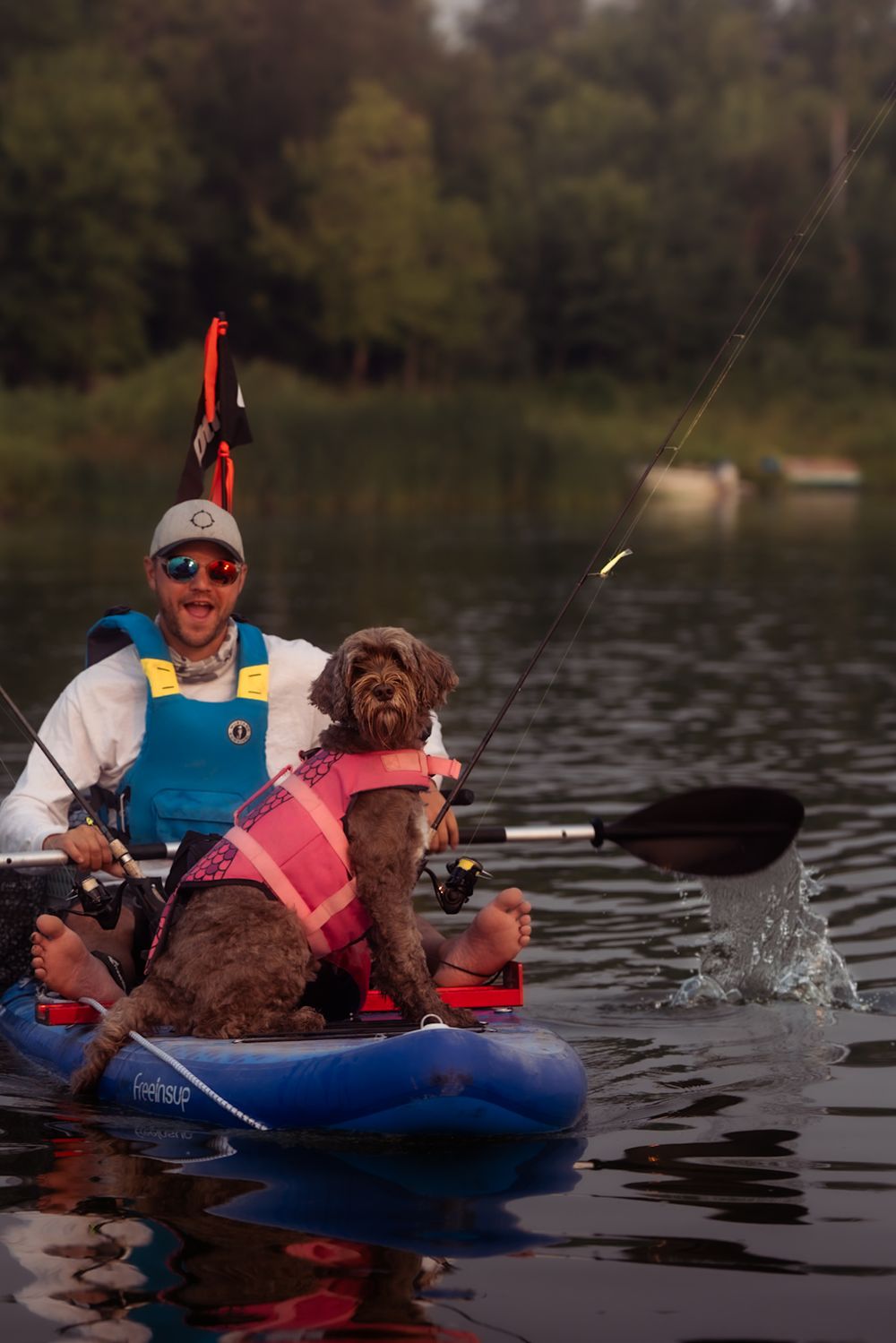 A chocolate brown dog wearing a life jacket joins companions in a blue kayak on a peaceful lake during an autumn day.