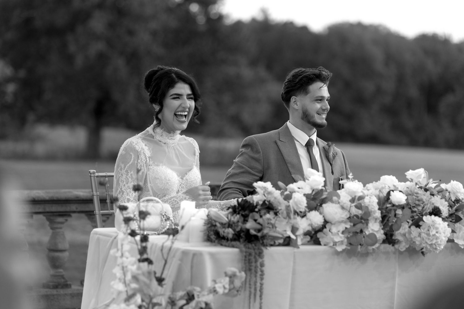 A black and white photo of a newlywed couple sitting at their reception table with floral arrangements.