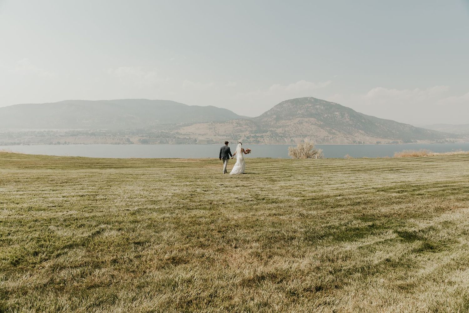 Bride and groom walking away across open field toward serene lake and rolling mountains in the distance.