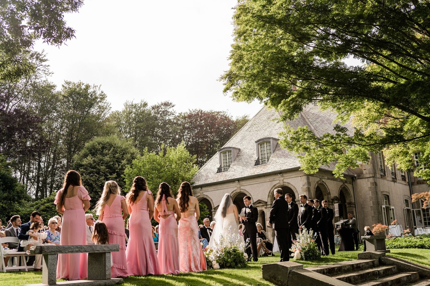 A wedding party walks across a lush green lawn in front of a white chapel on a sunny afternoon.
