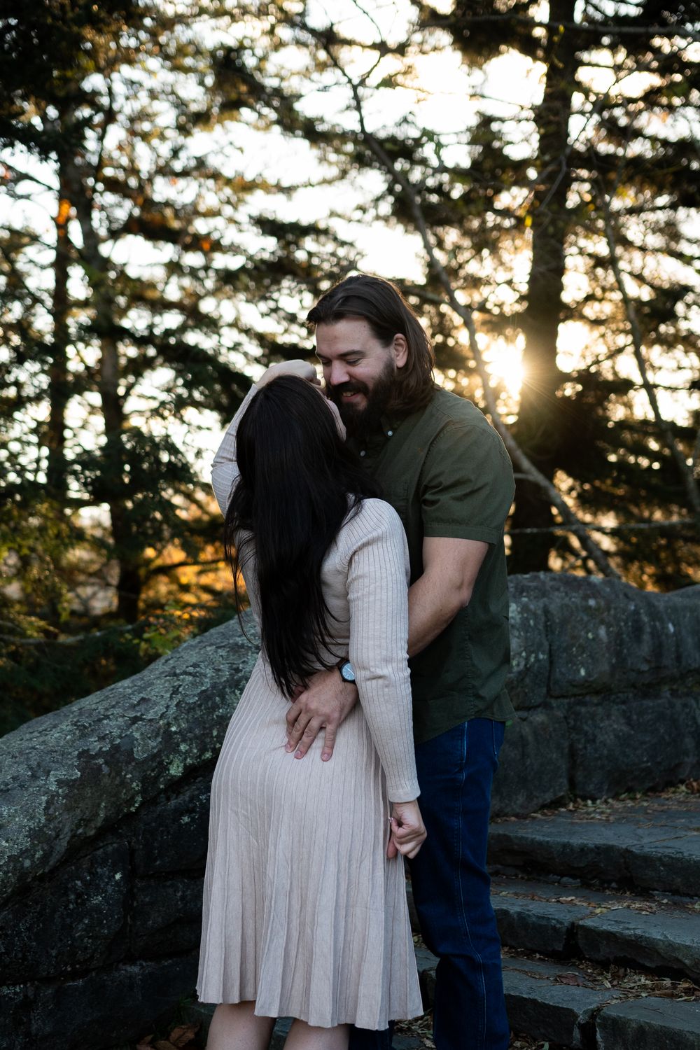 Romantic couple embraces on a stone wall during golden hour sunset in a forest setting.