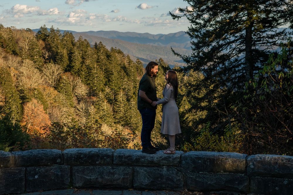 A couple shares a romantic moment at sunset on a stone wall overlooking mountain vistas and autumn forest scenery.