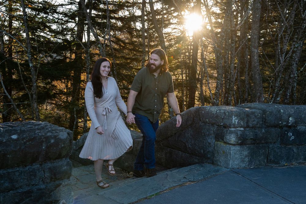 Intimate outdoor photoshoot with couple against sunlit evergreen trees and stone wall backdrop.