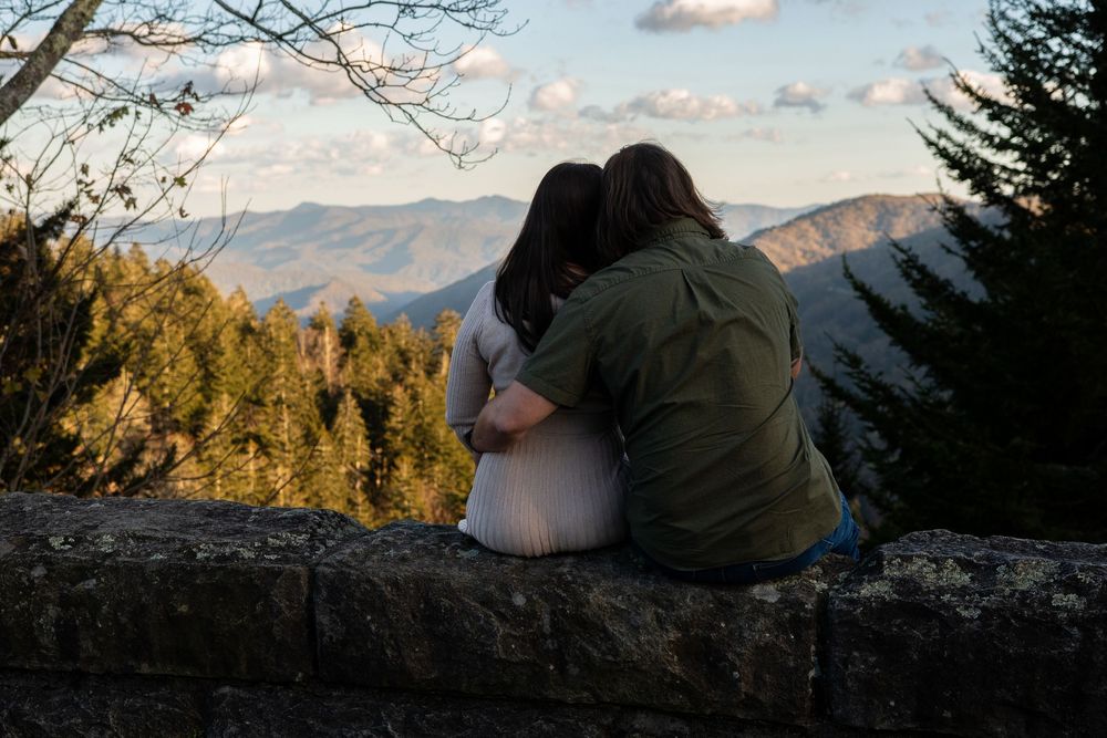 Couple enjoying a romantic mountain picnic at sunset with scenic views of rolling hills and evergreen trees.