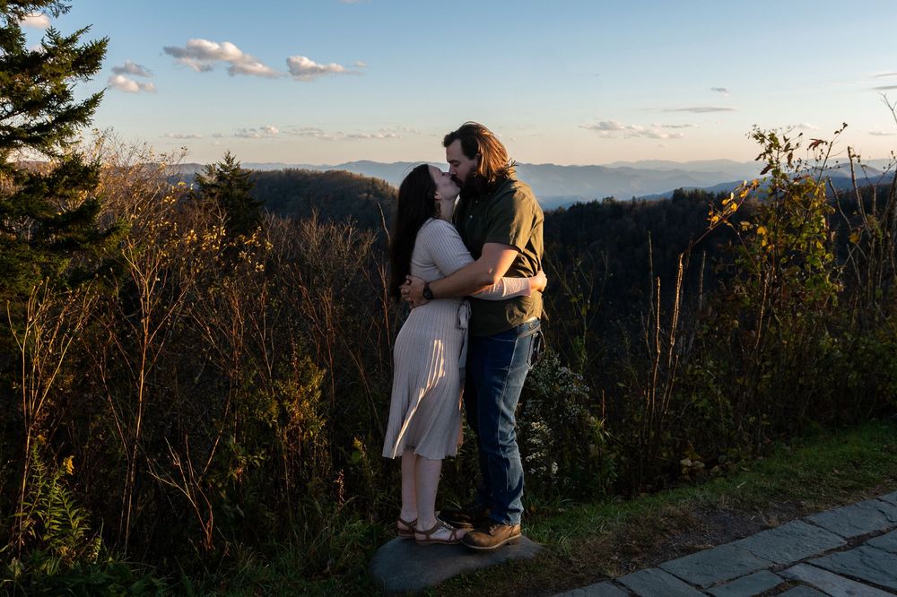 A couple shares a romantic moment at sunset on a scenic mountain overlook with rolling hills in the background.