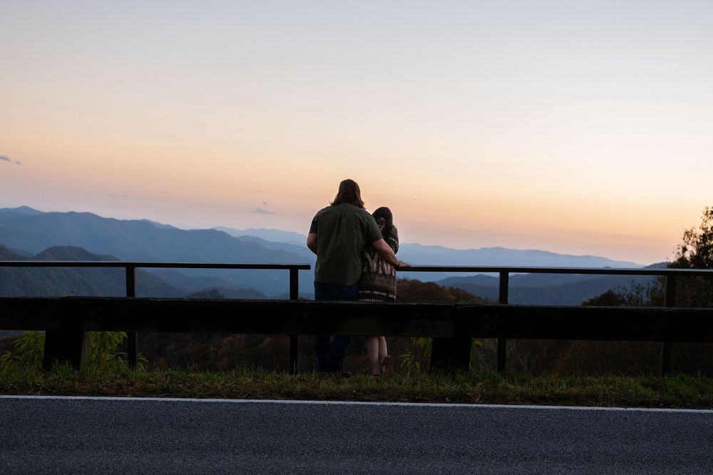 Silhouetted figures lean on railing overlooking mountain landscape during sunset.
