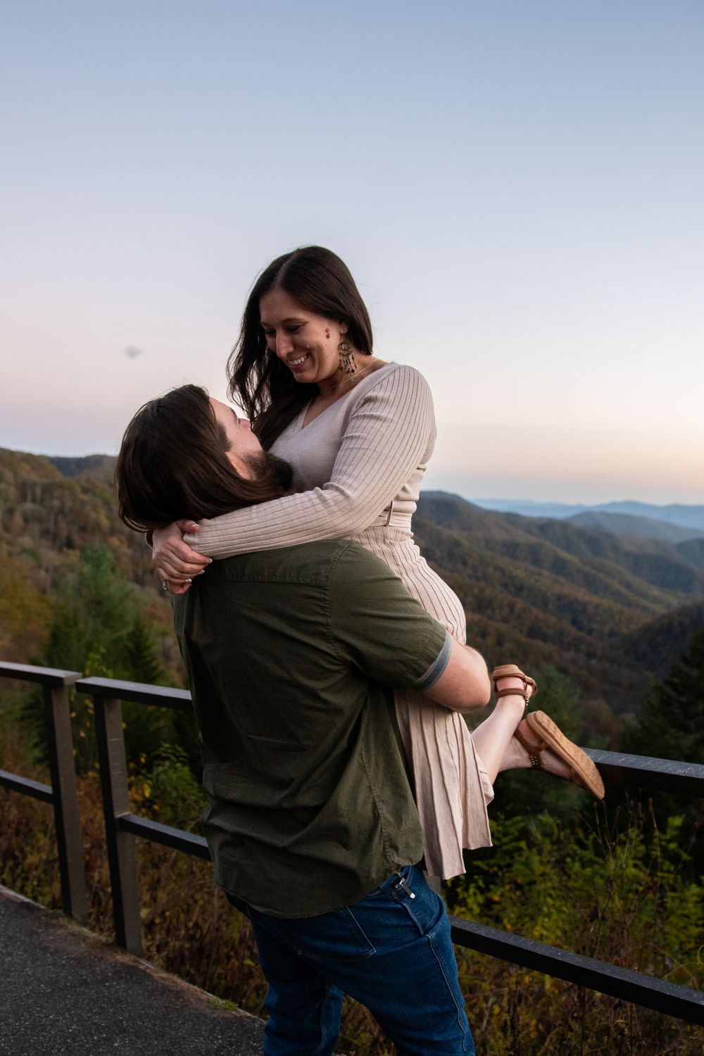 A romantic couple embraces on a scenic mountain overlook during sunset with rolling hills in the background.