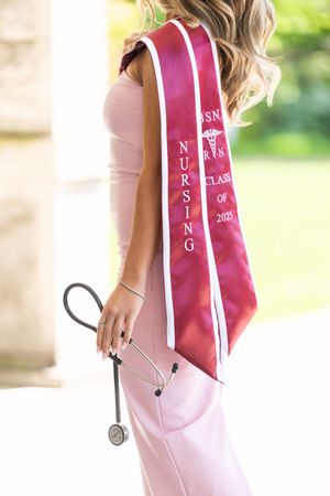 Graduate wearing pink sash and light dress holds stethoscope during nursing graduation.