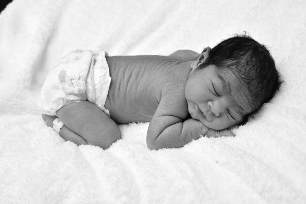 A sleeping baby lies on white bedding in a series of peaceful poses captured in black and white photography.