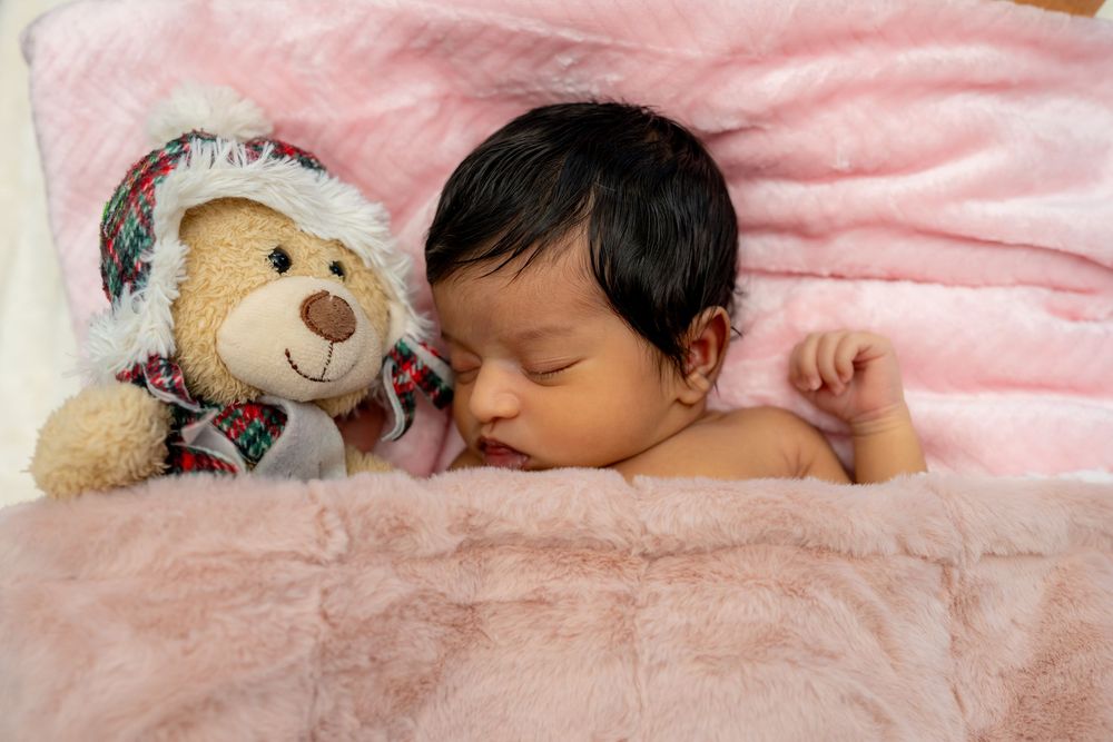 Sleeping baby lies peacefully on pink blanket next to plush teddy bear wearing winter hat and scarf.