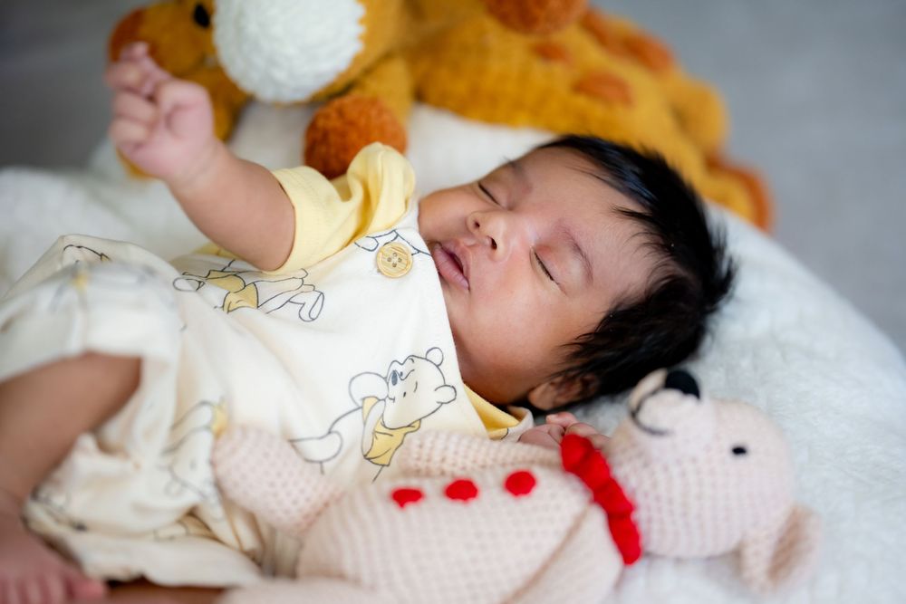 A sleeping baby in a cream-colored outfit lies on white bedding with a soft orange plush toy nearby.