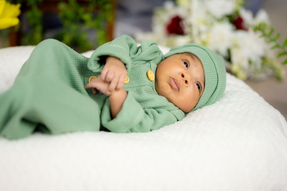 Newborn baby wearing soft mint green outfit and hat lying on white blanket with floral background.