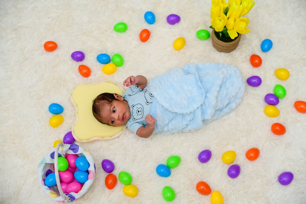 A baby lies on a white blanket surrounded by colorful plastic Easter eggs scattered on the floor.