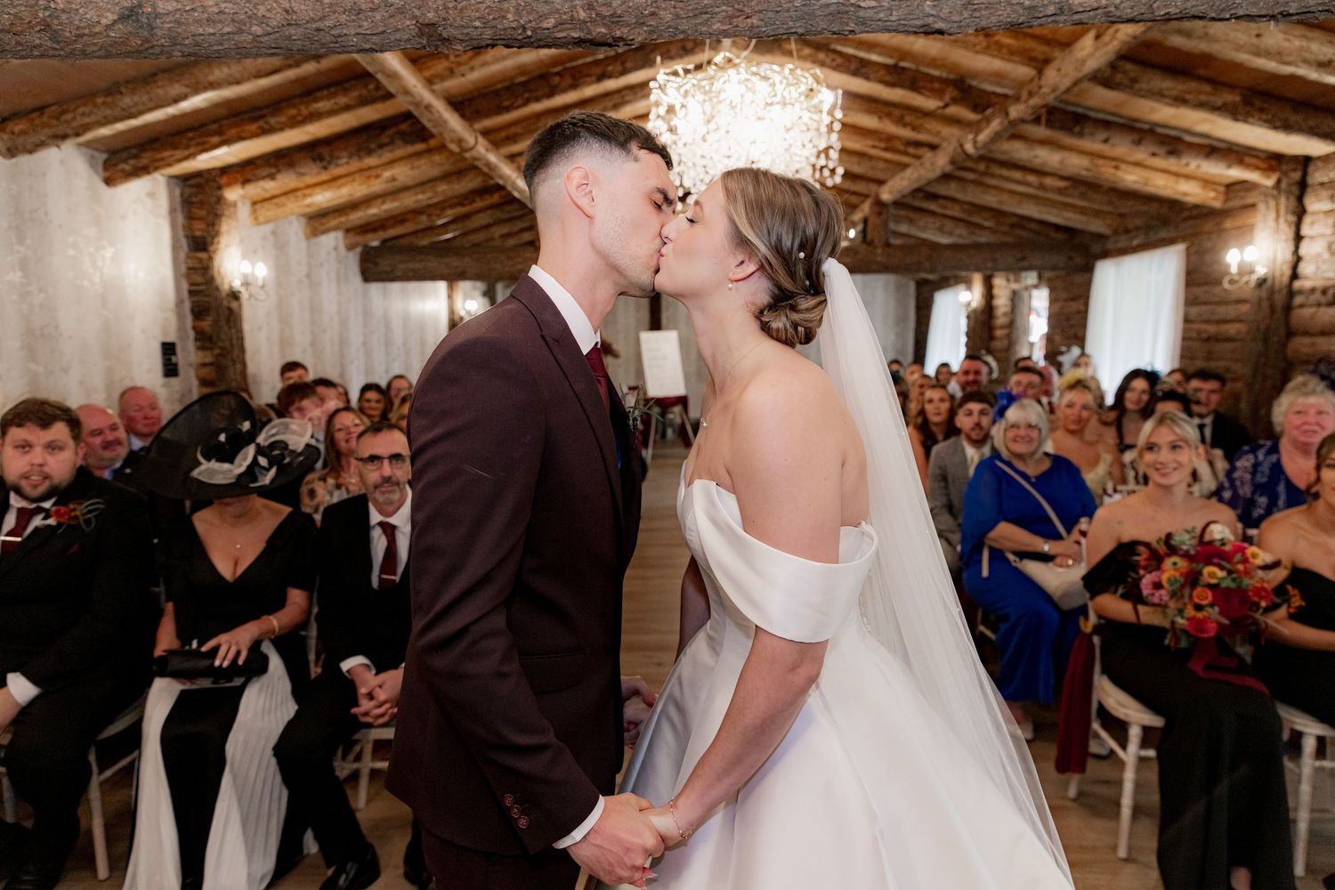 A romantic wedding ceremony takes place in a rustic venue with exposed wooden beams and gathered guests looking on.