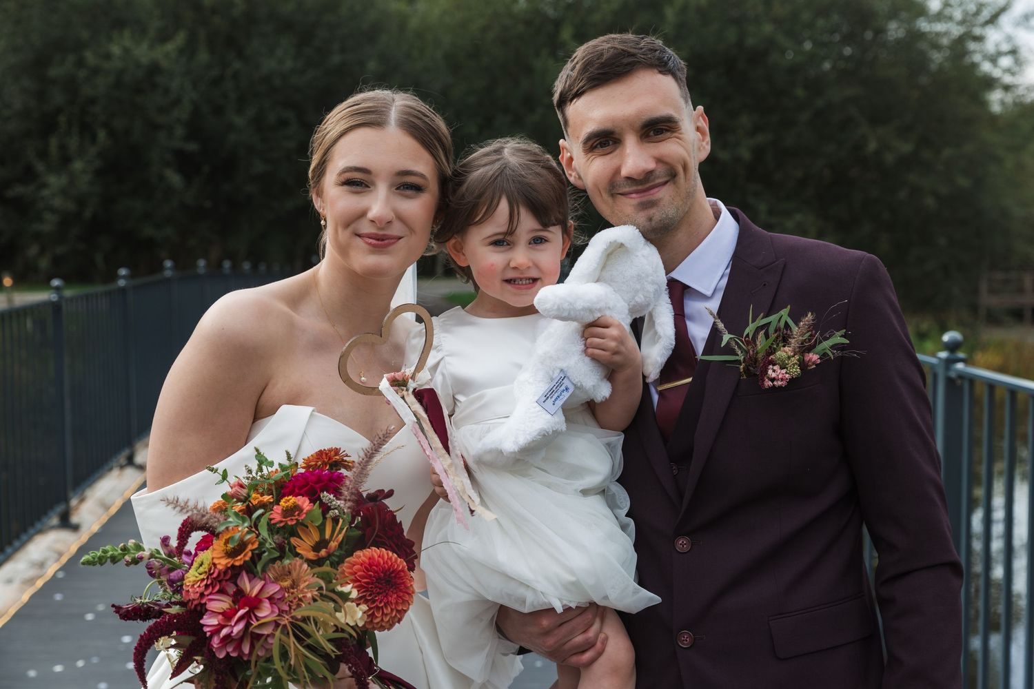 Wedding party poses outdoors in formal attire with burgundy and red floral bouquets.