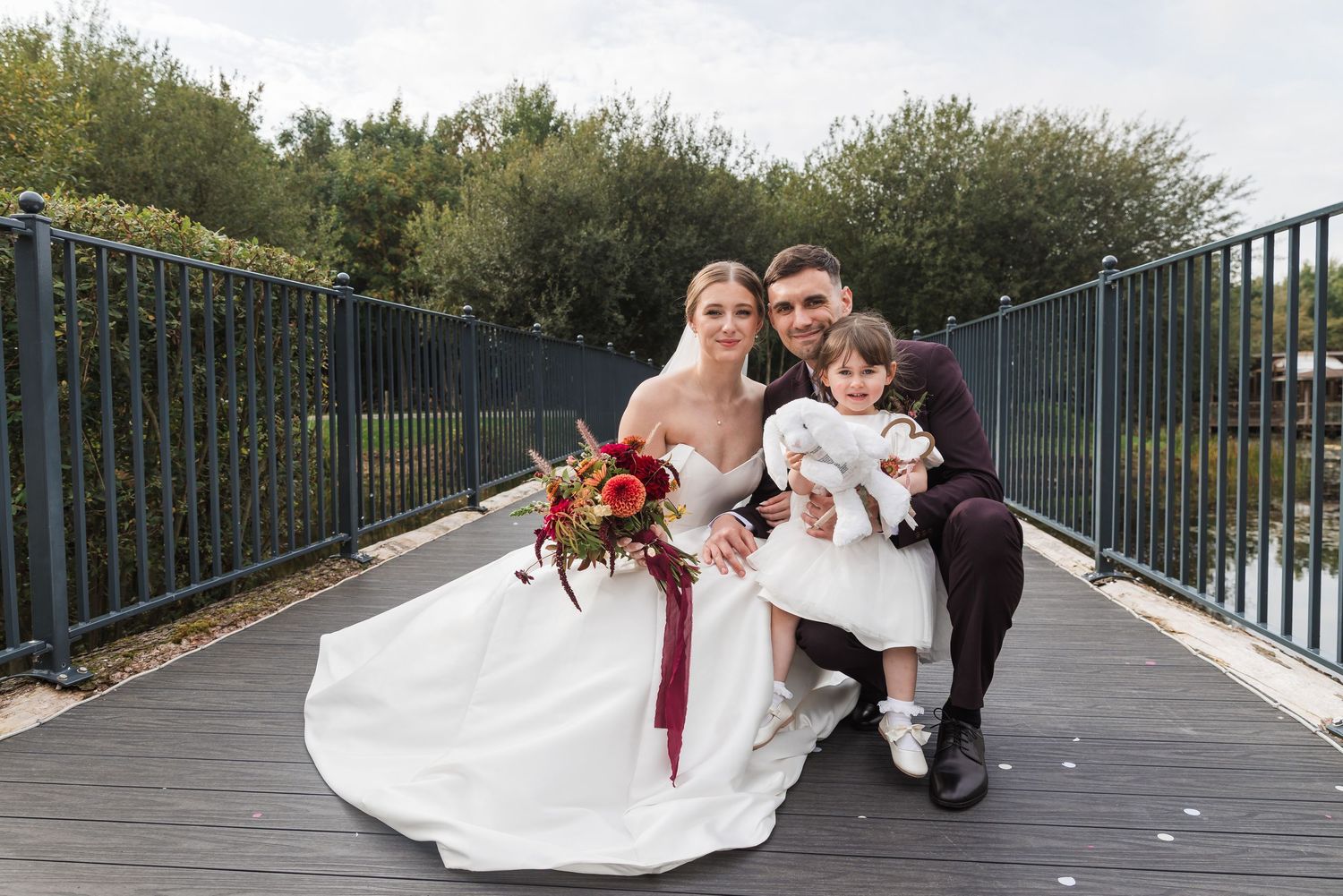 A couple in wedding attire poses on a wooden boardwalk with a red bouquet and white dress.
