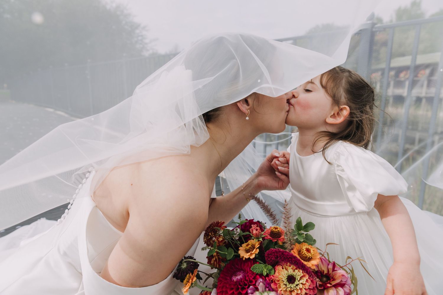 A bride in white dress holds a vibrant burgundy and pink floral bouquet during an outdoor wedding ceremony.