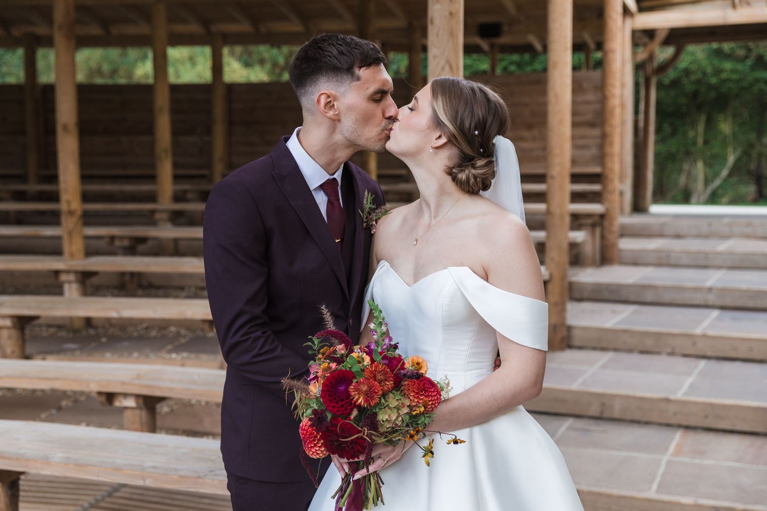 A romantic wedding couple shares a tender moment under a wooden pavilion with a vibrant red bouquet.