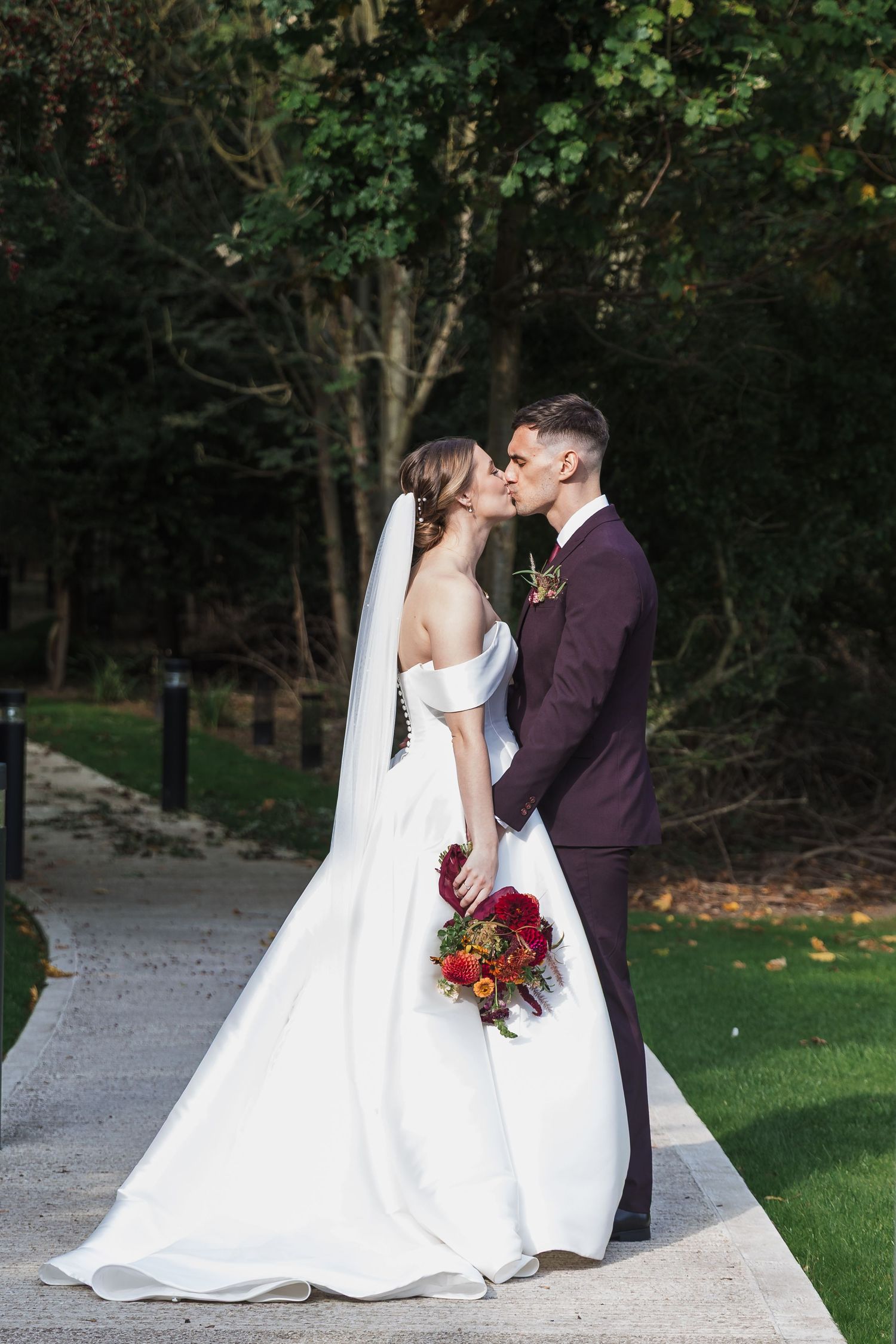A romantic wedding couple shares tender moments in a garden setting with a flowing white dress and burgundy suit.