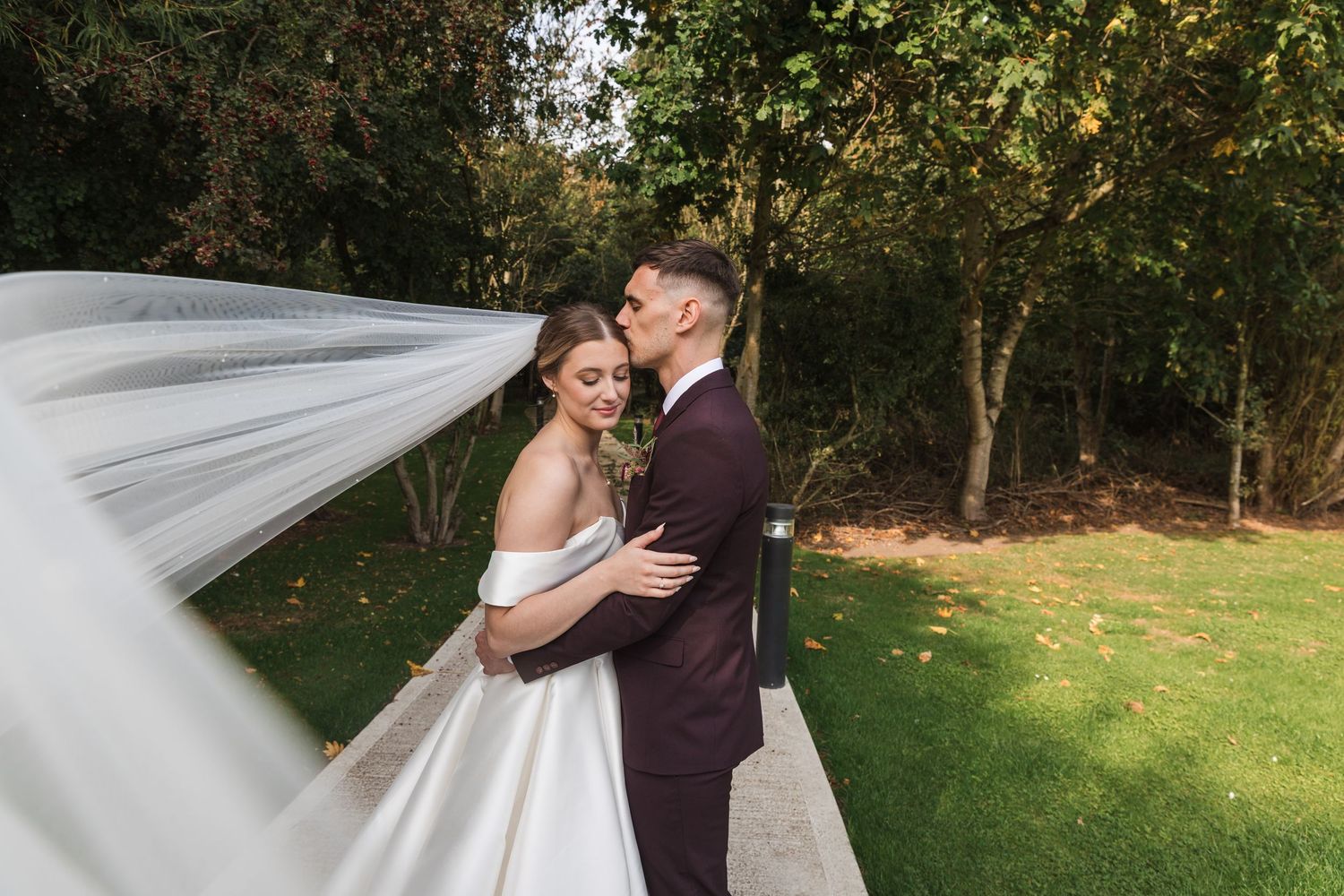 A romantic couple shares a kiss outdoors while a flowing white veil catches the breeze in a garden setting.