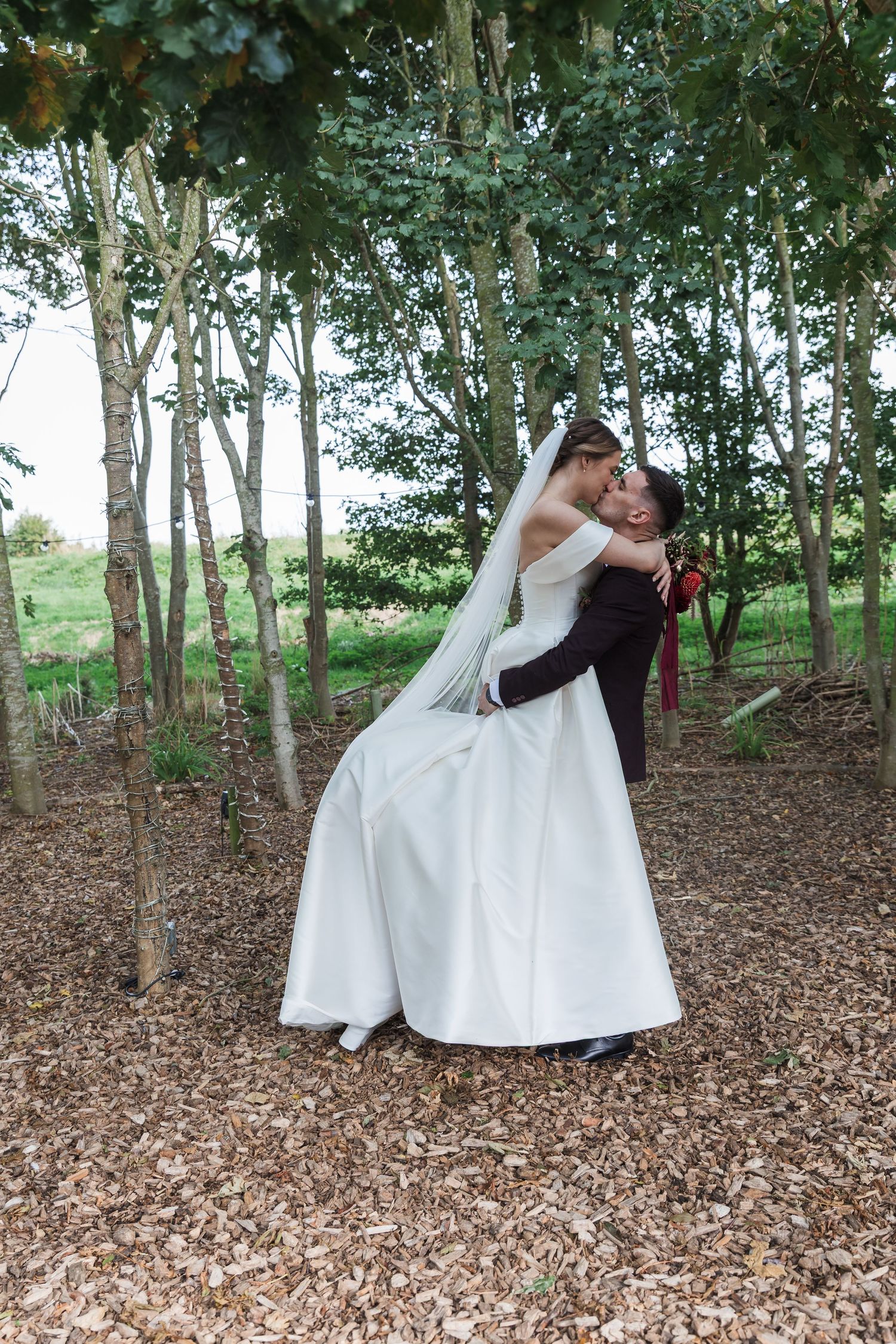 Wedding couple poses romantically in a wooded area with green trees and fallen leaves.
