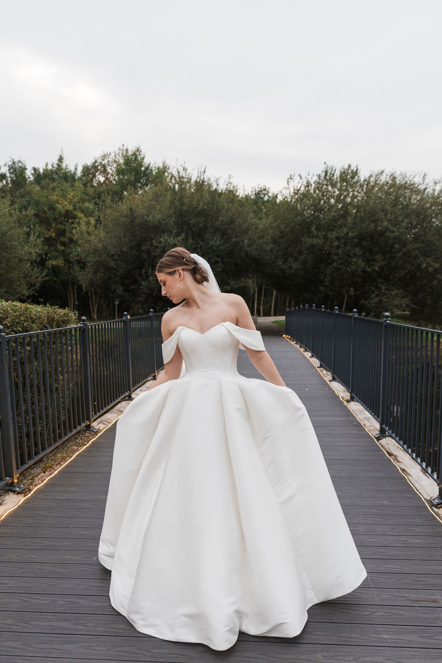A bride in an elegant off-shoulder white ballgown poses on a wooden bridge surrounded by greenery.