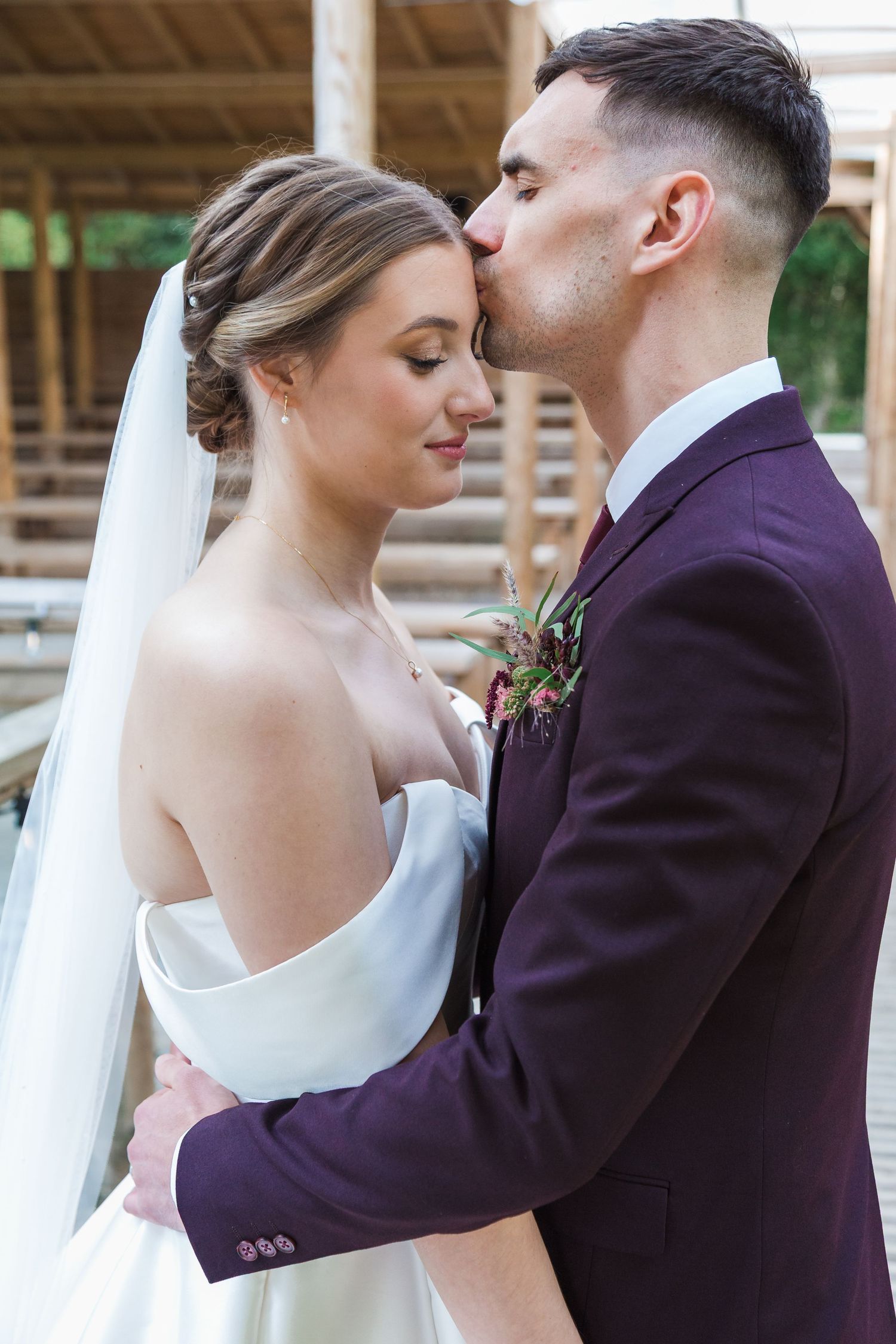 A tender moment between newlyweds as they embrace, the bride in white and groom in burgundy suit at an outdoor venue.