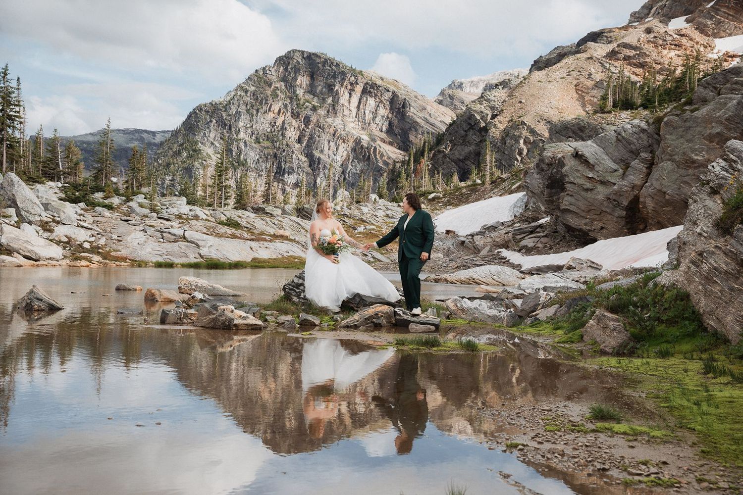 Two brides exchange vows beside a reflective alpine lake with snow-capped mountains.