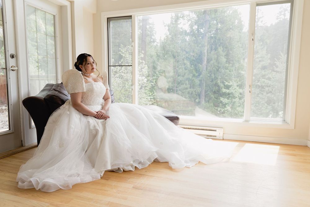 Bride in a flowing white ball gown sits gracefully by large windows with a lush green forest backdrop.