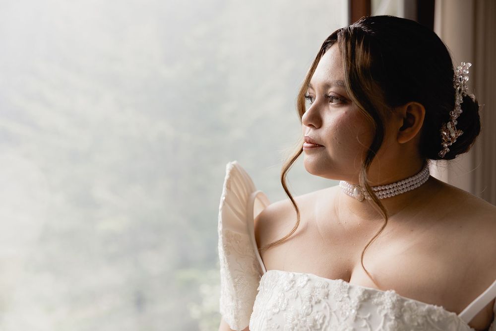 Elegant bride in white lace gown with pearl choker gazes thoughtfully out a window on her wedding day.